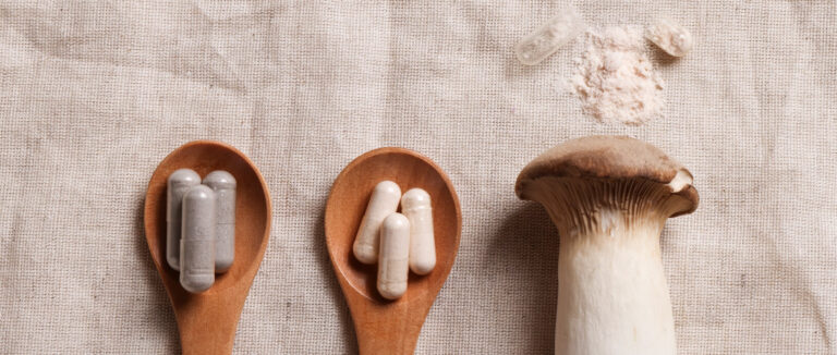 Two different kinds of mushroom extract capsules on wooden spoons. A raw mushroom and an open capsule with spilled powder are to the right.